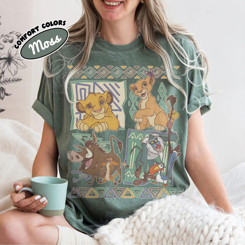 May include: A moss green t-shirt featuring a graphic print of characters from The Lion King, including Simba, Nala, Pumbaa, Timon, Rafiki, and Zazu, arranged in four panels with geometric borders and a Comfort Colors Moss label in the corner.