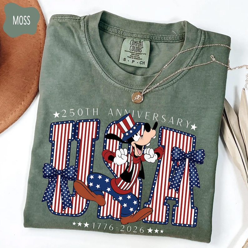 Może przedstawiać: Mchowo-zielony t-shirt z kr&oacute;tkim rękawem z grafiką psa z kresk&oacute;wki w patriotycznym cylindrze. Projekt zawiera napis 250TH ANNIVERSARY, USA oraz 1776-2026, z dużymi literami ozdobionymi czerwono-białymi paskami oraz niebieskimi gwiazdkami.