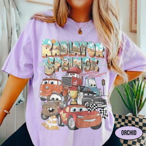 May include: A light purple t-shirt featuring a graphic print of Radiator Springs with Lightning McQueen and other characters. The design includes the text Radiator Springs and Lightning McQueen, a gold trophy, and a checkered flag in a vintage style.