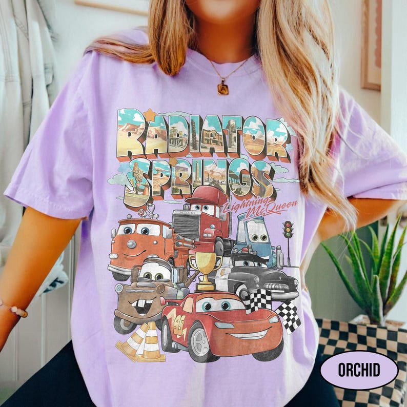 May include: A light purple t-shirt featuring a graphic print of Radiator Springs with Lightning McQueen and other characters. The design includes the text Radiator Springs and Lightning McQueen, a gold trophy, and a checkered flag in a vintage style.