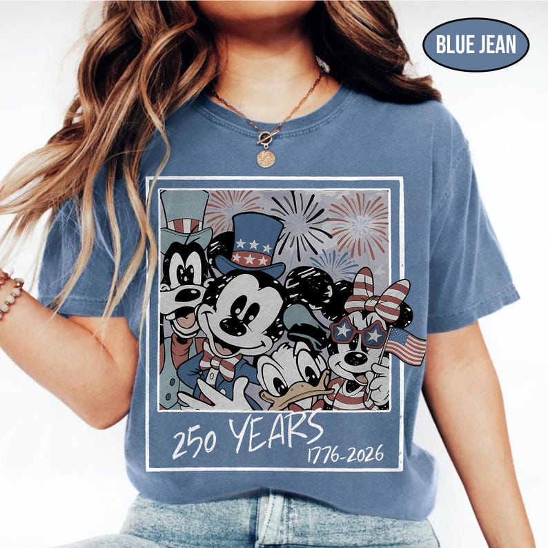 May include: A blue jean colored t-shirt featuring a graphic of cartoon characters wearing patriotic hats and accessories with fireworks in the background. The text 250 YEARS 1776-2026 is printed below the characters in a white, hand-drawn style font.