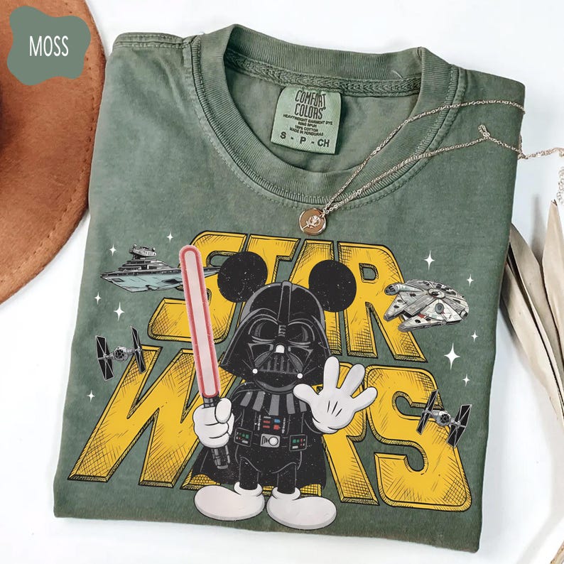 Puede incluir: Una camiseta verde musgo con un gr&aacute;fico de Darth Vader como Mickey Mouse sosteniendo un sable de luz, con las palabras "STAR WARS" en amarillo. La camiseta tambi&eacute;n incluye im&aacute;genes del Halc&oacute;n Milenario y otros elementos de Star Wars. Un collar plateado est&aacute; sobre la camiseta.