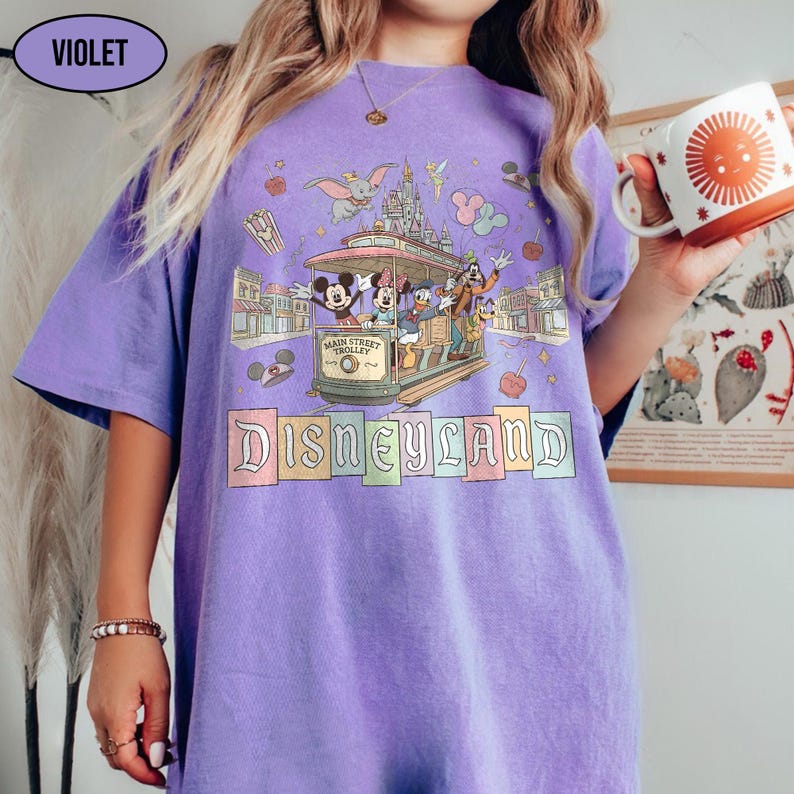 May include: A violet-colored t-shirt featuring a Disneyland design. The graphic includes a Main Street Trolley with Mickey, Minnie, Donald, and Goofy. The word "Disneyland" is displayed in colorful letters. A mug with a sun design is held in the hand.