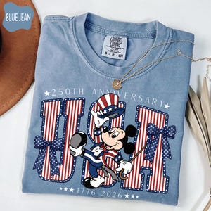 Może przedstawiać: Dżinsowo-niebieski t-shirt z grafiką Myszki Miki w patriotycznym cylindrze i garniturze. Wz&oacute;r zawiera napis 250TH ANNIVERSARY oraz 1776-2026, a także duże litery USA w czerwono-białe paski oraz niebieskie kokardy w białe kropki.