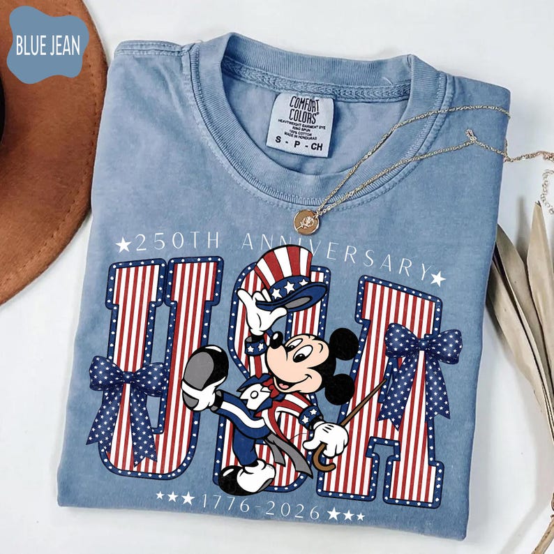 Może przedstawiać: Dżinsowo-niebieski t-shirt z grafiką Myszki Miki w patriotycznym cylindrze i garniturze. Wz&oacute;r zawiera napis 250TH ANNIVERSARY oraz 1776-2026, a także duże litery USA w czerwono-białe paski oraz niebieskie kokardy w białe kropki.