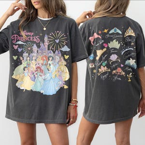 K&ouml;nnte beinhalten: Ein anthrazitfarbenes Oversize-T-Shirt mit einem Frontdruck von Disney-Prinzessinnen vor einem Schloss mit Feuerwerk und dem Text Disney Princess. Die R&uuml;ckseite zeigt verschiedene Kronen und Accessoires mit Namen.