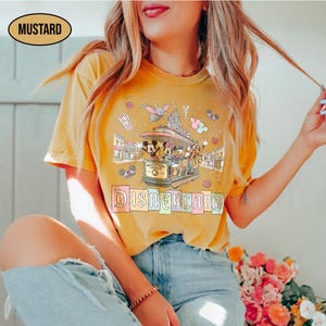 May include: Mustard-colored t-shirt featuring a Disneyland design. The graphic includes a trolley with Mickey Mouse, Minnie Mouse, Donald Duck, and Goofy. The word "Disneyland" is written in pastel colors below the trolley.