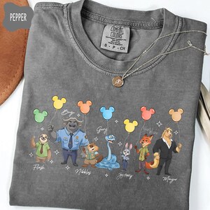 Puede incluir: Una camiseta gris Comfort Colors con personajes de dibujos animados de Zootopia sosteniendo globos con forma de Mickey Mouse. Los personajes incluyen a Flash, Bogo, Gary, Nibbles, Judy Hopps, Nick Wilde y Mayor. Tambi&eacute;n se ve un collar plateado con un colgante.