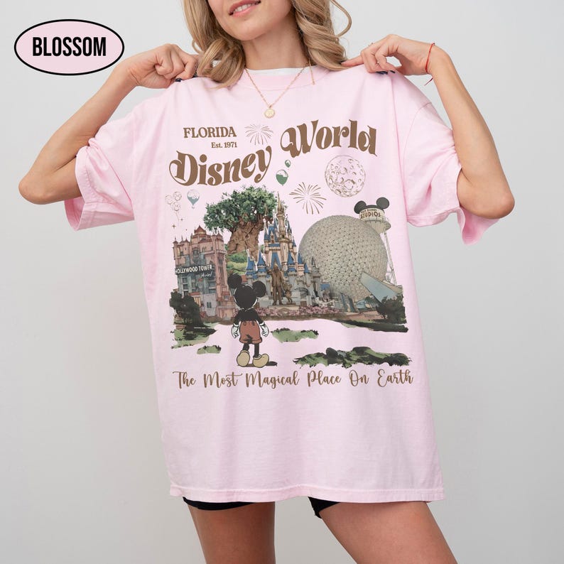 Puede incluir: Camiseta rosa claro con un gr&aacute;fico de Disney World. El dise&ntilde;o incluye el texto "Florida Est. 1971 Disney World" y una ilustraci&oacute;n de Mickey Mouse frente a los puntos de referencia del parque. Tambi&eacute;n se incluye el texto "The Most Magical Place On Earth".