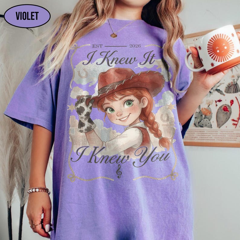 May include: A violet t-shirt featuring a graphic of a cartoon cowgirl wearing a brown hat and a cow-print glove. The text reads EST 2026, I Knew It, and I Knew You, accompanied by a treble clef symbol. The design includes clouds and stars.