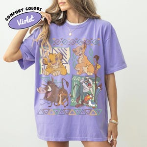 May include: A violet short-sleeve t-shirt featuring a graphic print of animated lion characters, a warthog, a meerkat, a mandrill, and a hornbill. The design includes geometric patterns and the text COMFORT COLORS Violet in the upper left corner.