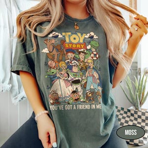 May include: A moss green t-shirt featuring a colorful graphic of characters from the animated film "Toy Story." The design includes the words "TOY STORY" and "YOU'VE GOT A FRIEND IN ME."