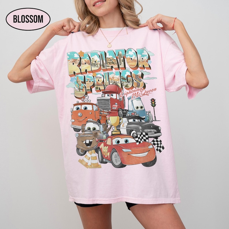 May include: A light pink t-shirt featuring a graphic print of Radiator Springs with characters from the movie Cars, including Lightning McQueen, Mater, and others. The text reads Radiator Springs, Lightning McQueen, and Blossom.