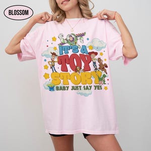 K&ouml;nnte beinhalten: Ein hellrosa T-Shirt mit einem Grafikdruck von Toy Story-Figuren, darunter Buzz Lightyear, Woody, Jessie, Bullseye, Slinky Dog, Rex und ein Alien. Der Text lautet IT'S A TOY STORY BABY JUST SAY YES in gelber und roter Schrift.