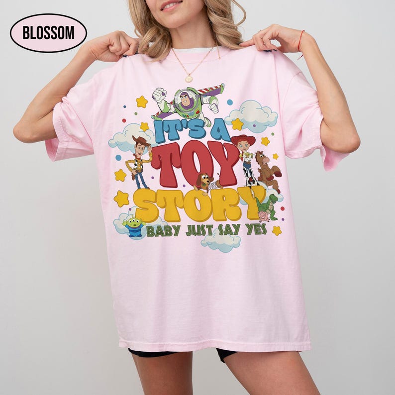 K&ouml;nnte beinhalten: Ein hellrosa T-Shirt mit einem Grafikdruck von Toy Story-Figuren, darunter Buzz Lightyear, Woody, Jessie, Bullseye, Slinky Dog, Rex und ein Alien. Der Text lautet IT'S A TOY STORY BABY JUST SAY YES in gelber und roter Schrift.