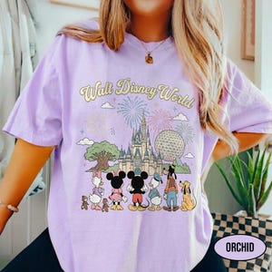 May include: A lilac t-shirt featuring a graphic of the Walt Disney World castle, fireworks, and Disney characters. The text "Walt Disney World" is arched above the image. The word "ORCHID" is in a white oval at the bottom right.