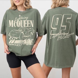 Puede incluir: Camiseta verde oliva con un dise&ntilde;o de Lightning McQueen. La parte delantera presenta un dise&ntilde;o de coche de dibujos animados con el texto "Lightning McQueen", "95" y "Think Fast". La parte trasera muestra un gran "95" y el texto "Lightning McQueen".