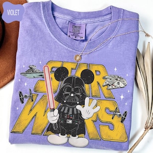 Puede incluir: Camiseta violeta Comfort Colors con un gr&aacute;fico de Star Wars. El dise&ntilde;o incluye a Darth Vader con orejas de Mickey Mouse, un sable de luz y el texto "STAR WARS" en amarillo. Un collar dorado est&aacute; sobre la camiseta.
