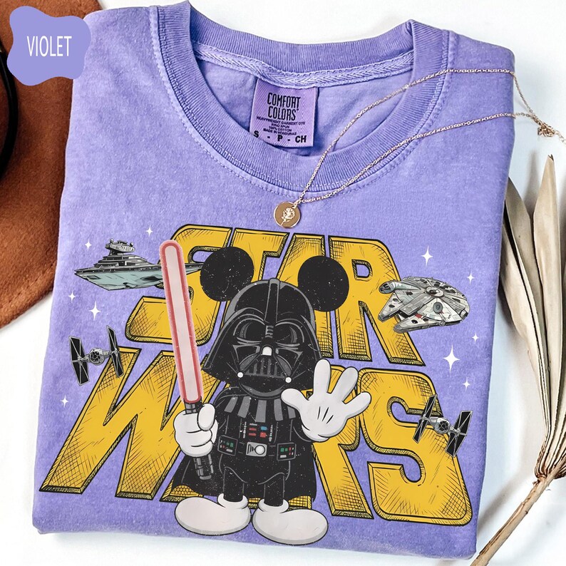 Puede incluir: Camiseta violeta Comfort Colors con un gr&aacute;fico de Star Wars. El dise&ntilde;o incluye a Darth Vader con orejas de Mickey Mouse, un sable de luz y el texto "STAR WARS" en amarillo. Un collar dorado est&aacute; sobre la camiseta.