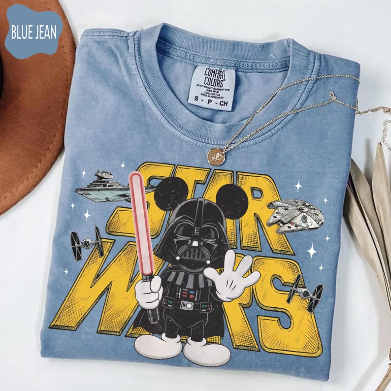 Puede incluir: Una camiseta azul Comfort Colors con un personaje de dibujos animados de Darth Vader con orejas de Mickey Mouse sosteniendo un sable de luz. La camiseta tiene las palabras "STAR WARS" en amarillo, con im&aacute;genes del Halc&oacute;n Milenario y otras naves espaciales.