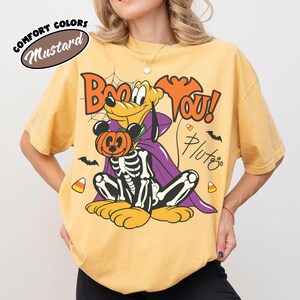 May include: A mustard yellow t-shirt featuring a graphic of Pluto wearing a purple cape and a skeleton costume, holding a Mickey Mouse pumpkin. The text reads Boo YOU! and Pluto. A Comfort Colors Mustard label is visible in the upper left corner.