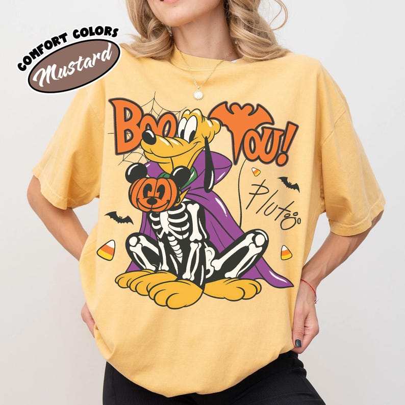 May include: A mustard yellow t-shirt featuring a graphic of Pluto wearing a purple cape and a skeleton costume, holding a Mickey Mouse pumpkin. The text reads Boo YOU! and Pluto. A Comfort Colors Mustard label is visible in the upper left corner.