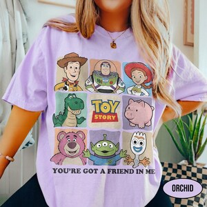 May include: A light purple t-shirt featuring a grid of Toy Story characters, including Woody, Buzz Lightyear, and Jessie. The words "TOY STORY" are in the center, with "YOU'RE GOT A FRIEND IN ME" below. The word "ORCHID" is in a white oval.