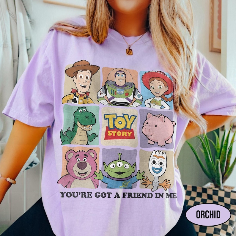 May include: A light purple t-shirt featuring a grid of Toy Story characters, including Woody, Buzz Lightyear, and Jessie. The words "TOY STORY" are in the center, with "YOU'RE GOT A FRIEND IN ME" below. The word "ORCHID" is in a white oval.