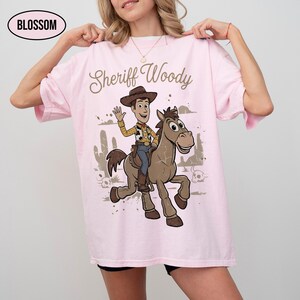 May include: A light pink t-shirt featuring a graphic of Sheriff Woody riding a brown horse. The text "Sheriff Woody" is printed above the character in a brown script font. A small oval logo in the top left corner contains the text "BLOSSOM".