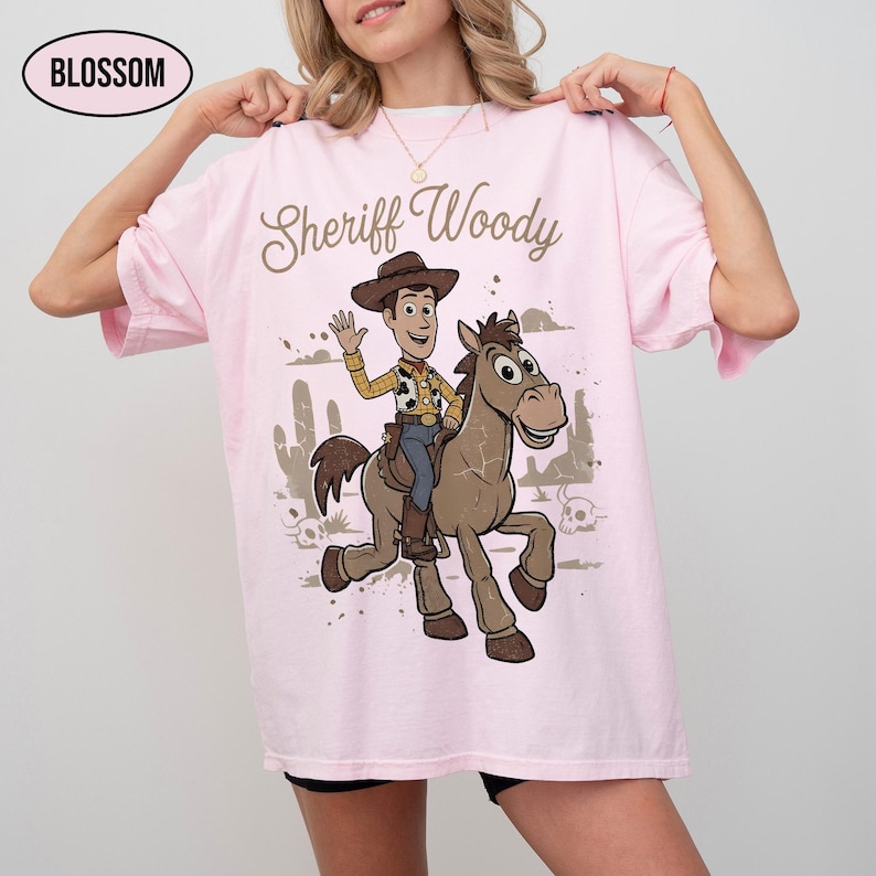 May include: A light pink t-shirt featuring a graphic of Sheriff Woody riding a brown horse. The text "Sheriff Woody" is printed above the character in a brown script font. A small oval logo in the top left corner contains the text "BLOSSOM".