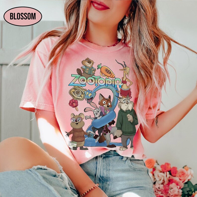 May include: Pink t-shirt featuring the word "Zootopia" in a colorful design, along with cartoon characters from the movie. The shirt has a crew neck and short sleeves. The word "Blossom" is in a white oval in the upper left corner.