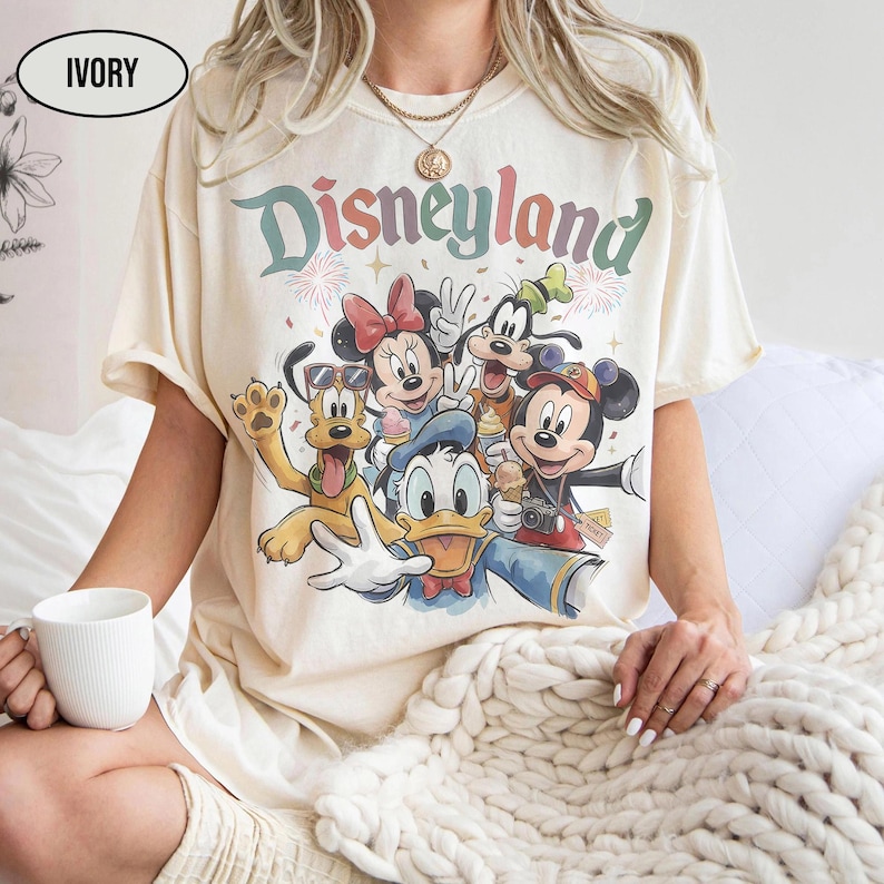 May include: Ivory-colored t-shirt featuring a colorful graphic of Disney characters, including Mickey Mouse, Minnie Mouse, Donald Duck, Goofy, and Pluto. The word "Disneyland" is printed above the characters in a rainbow font. The shirt has short sleeves.