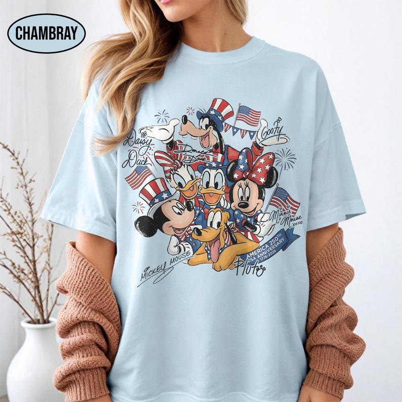 Może przedstawiać: Jasnoniebieska koszulka z grafiką postaci Disneya Myszki Miki, Myszki Minnie, Kaczora Donalda, Kaczki Daisy, Goofy'ego i Pluto, z amerykańskimi flagami i patriotycznymi wzorami. Koszulka ma napis "America 250th Anniversary 1776-2026".