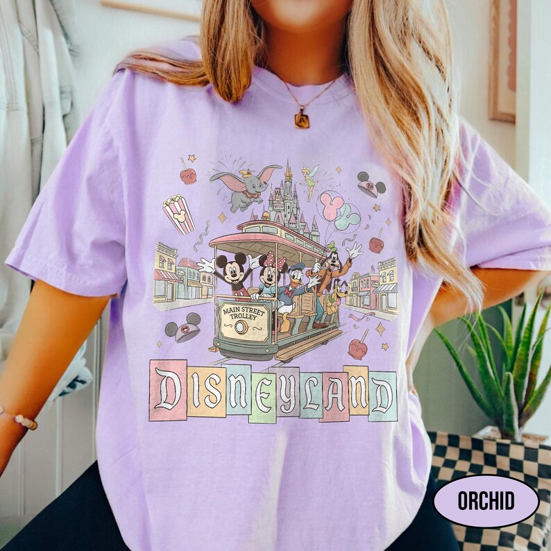 May include: A light purple t-shirt featuring a Disneyland design. The graphic includes a trolley with Mickey and friends, a castle, and the word "Disneyland" in a colorful, vintage font. The shirt has short sleeves.