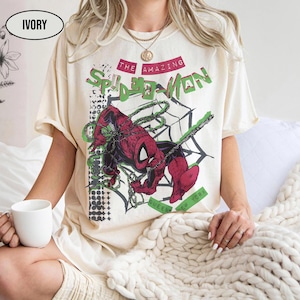 May include: Ivory-colored t-shirt featuring a graphic of Spider-Man in a web. The design includes the text "THE AMAZING SPIDER-MAN" in red and green, with "QUEENS NEW YORK" below. The shirt is short-sleeved.
