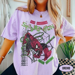 May include: A light purple t-shirt featuring a graphic of Spider-Man in a web. The design includes the text "THE AMAZING SPIDER-MAN" in red and green, with "QUEENS NEW YORK" below. The shirt is a soft, comfortable material.