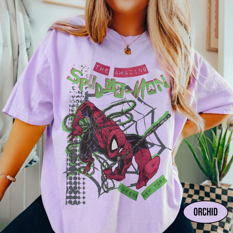 May include: A light purple t-shirt featuring a graphic of Spider-Man in a web. The design includes the text "THE AMAZING SPIDER-MAN" in red and green, with "QUEENS NEW YORK" below. The shirt is a soft, comfortable material.