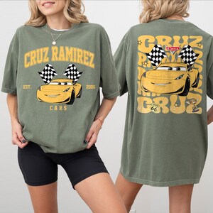 Peut inclure: T-shirt vert olive avec un dessin de voiture jaune et le texte "CRUZ RAMIREZ". Le devant du t-shirt porte l'inscription "EST. 2006 CARS". Le dos du t-shirt pr&eacute;sente le m&ecirc;me motif de voiture et le m&ecirc;me texte.