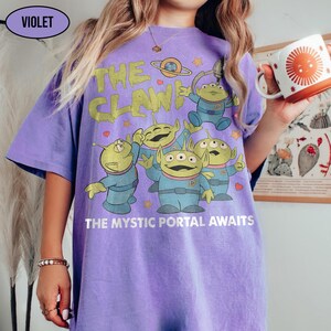 May include: Violet-colored t-shirt featuring a graphic of green aliens with the text "THE CLAW!" and "THE MYSTIC PORTAL AWAITS" in yellow and white. The shirt has a relaxed fit.