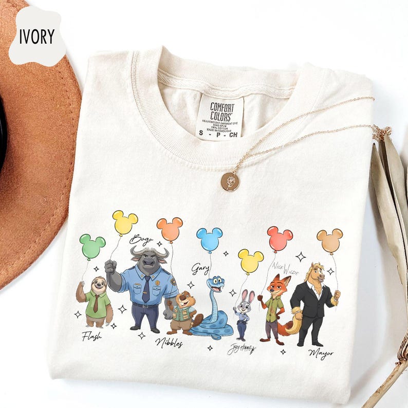 Puede incluir: Camiseta de color marfil con personajes de dibujos animados sosteniendo globos con forma de Mickey Mouse. Los personajes incluyen Flash, Bogo, Nibbles, Gary, Judy Hopps, Nick Wilde y Mayor. La camiseta tiene una etiqueta Comfort Colors y un collar dorado.