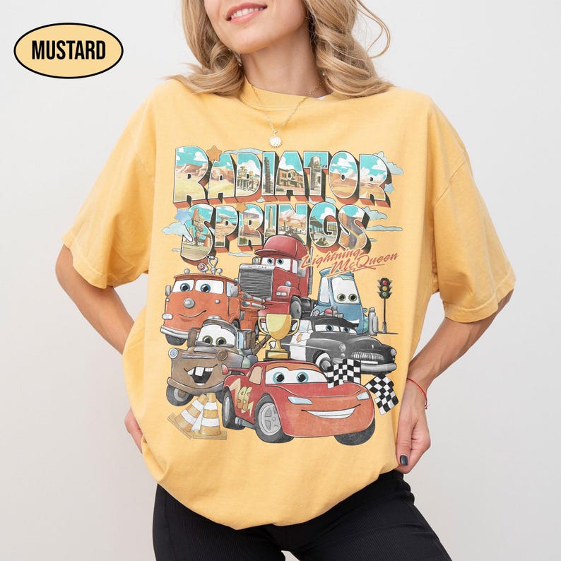 May include: A mustard yellow short-sleeve t-shirt featuring a graphic print of Radiator Springs and Lightning McQueen from the movie Cars. The design includes various character vehicles, a gold trophy, and checkered flags with the text Radiator Springs.