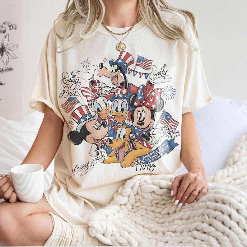 Może przedstawiać: Kremowa koszulka z grafiką postaci Disneya: Myszki Miki, Myszki Minnie, Kaczora Donalda, Kaczki Daisy, Goofy'ego i Plutona, z amerykańskimi flagami i patriotycznymi wzorami. Koszulka zawiera napis "America 250th USA Anniversary 1776-2026".
