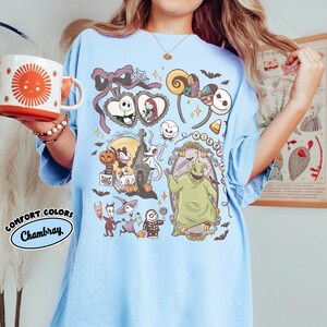 May include: Light blue t-shirt featuring a colorful graphic of characters from The Nightmare Before Christmas. The design includes Jack Skellington, Sally, Oogie Boogie, and Zero. The shirt has the words "Comfort Colors Chambray" on the bottom left.