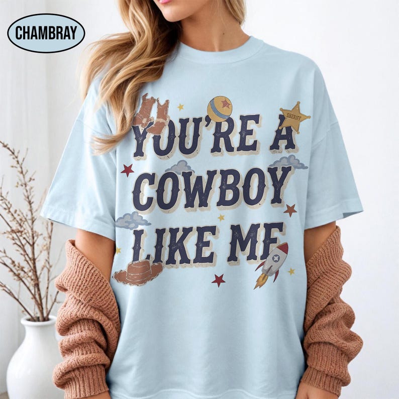 Puede incluir: Una camiseta azul claro con el texto azul oscuro "YOU'RE A COWBOY LIKE ME" rodeado de ilustraciones de botas de vaquero, una pelota, una estrella de sheriff, nubes, un sombrero de vaquero, un cohete y peque&ntilde;as estrellas rojas y amarillas.