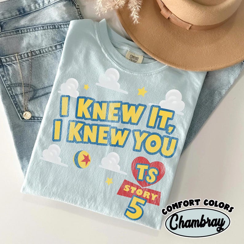 Puede incluir: Una camiseta azul claro con el texto amarillo I KNEW IT, I KNEW YOU, nubes blancas, estrellas amarillas, un coraz&oacute;n rojo con las letras TS y el texto STORY 5. La camiseta tiene cuello redondo y una etiqueta de la marca Comfort Colors.