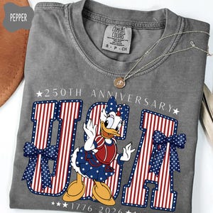 Może przedstawiać: Szary t-shirt z kr&oacute;tkim rękawem z grafiką Daisy Duck w patriotycznym stroju. Napis 250TH ANNIVERSARY oraz 1776-2026 otacza duży napis USA, wypełniony czerwono-białymi paskami oraz niebieskimi kokardami w białe kropki.