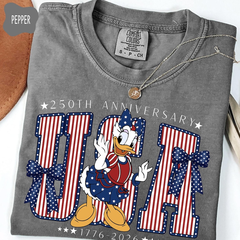 Może przedstawiać: Szary t-shirt z kr&oacute;tkim rękawem z grafiką Daisy Duck w patriotycznym stroju. Napis 250TH ANNIVERSARY oraz 1776-2026 otacza duży napis USA, wypełniony czerwono-białymi paskami oraz niebieskimi kokardami w białe kropki.