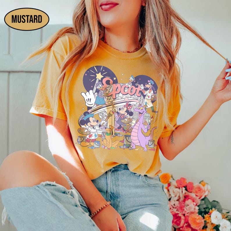 May include: Mustard-colored t-shirt featuring a colorful graphic of Disney characters, including Mickey Mouse, Minnie Mouse, and Figment, with the word "Epcot" in a stylized font. The shirt has a crew neck and short sleeves.