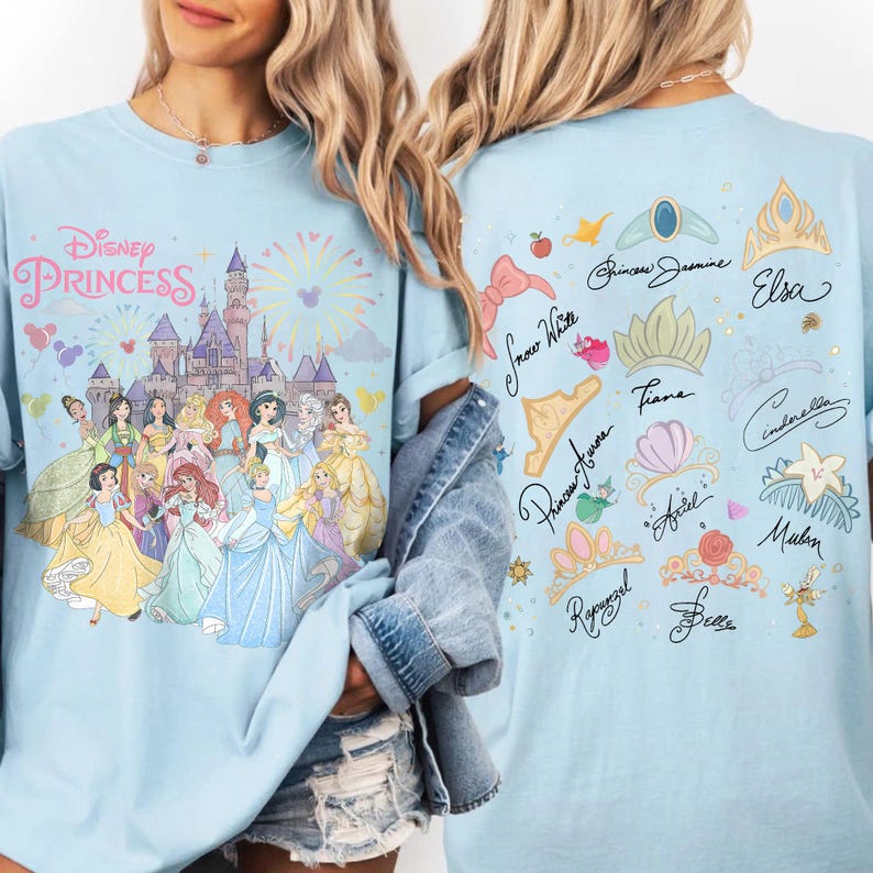 K&ouml;nnte beinhalten: Ein hellblaues T-Shirt mit einer Disney-Prinzessinnen-Grafik auf der Vorderseite, die ein Schloss und verschiedene Figuren zeigt. Die R&uuml;ckseite zeigt eine Sammlung illustrierter Kronen und Accessoires mit den Namen verschiedener Disney-Prinzessinnen.
