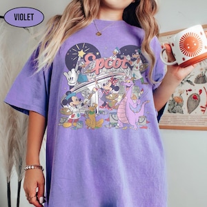 May include: A violet-colored t-shirt featuring a graphic of Disney characters painting the word "Epcot" on a bridge. The design includes Mickey Mouse, Minnie Mouse, Donald Duck, and a purple dragon. The shirt has short sleeves and a crew neck.